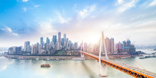 A panoramic view of a modern city skyline with numerous high-rise buildings under a bright, partly cloudy sky. In the foreground, an orange and white suspension bridge spans across a wide river, leading towards the city situated on both sides of the